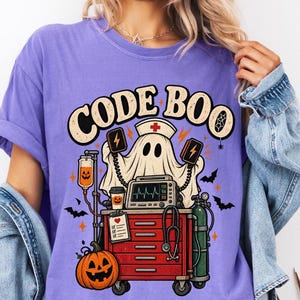 May include: A violet t-shirt featuring a graphic print with the text CODE BOO. The design shows a ghost wearing a nurse cap, a red medical cart, a heart rate monitor, a stethoscope, an IV bag with a pumpkin, and a large orange pumpkin.