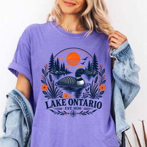 May include: A violet short-sleeve t-shirt featuring a graphic of a loon swimming on a lake with pine trees, cattails, and an orange sun. The text reads Lake Ontario Est. 1656, framed by a circular design with floral accents and a compass icon.