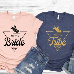 May include: Two t-shirts with a graphic design of a cowboy riding a bull. The pink shirt says "Austin Bride TX" and the blue shirt says "Austin Tribe TX".