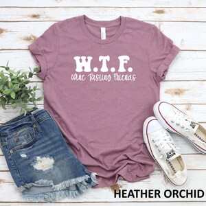K&ouml;nnte beinhalten: Ein T-Shirt in der Farbe Heide-Orchidee mit dem Text "W.T.F. wine Tasting Friends" in Wei&szlig;. Das Shirt liegt flach auf einem wei&szlig;en Holzgrund mit einem Paar wei&szlig;en Canvas-Schuhen und einer Jeans-Shorts.