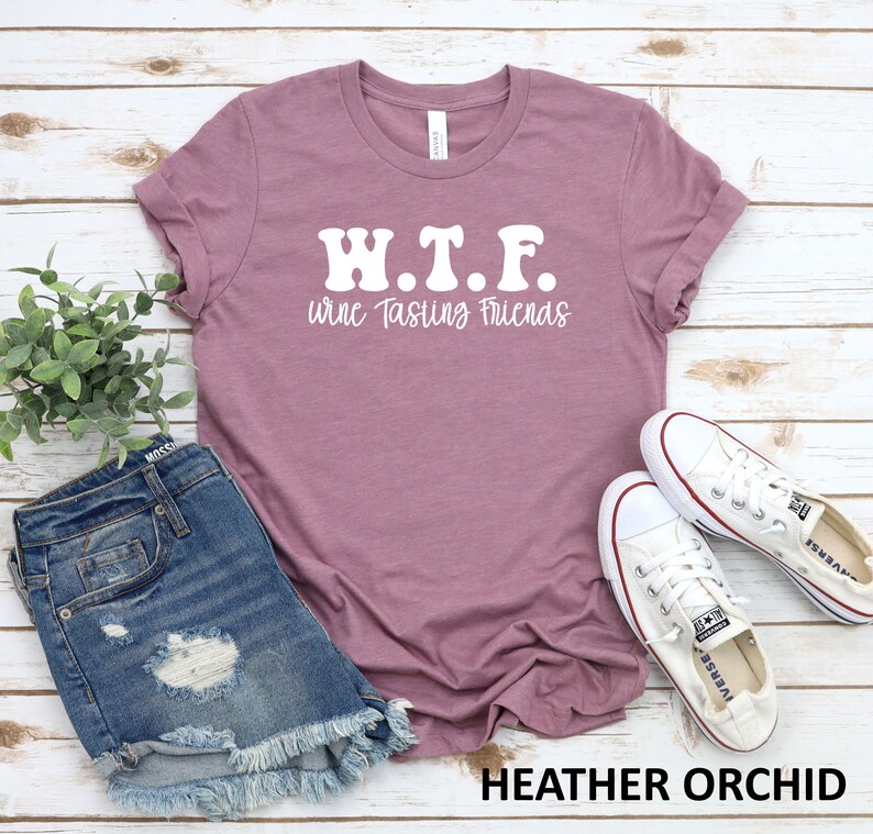 K&ouml;nnte beinhalten: Ein T-Shirt in der Farbe Heide-Orchidee mit dem Text "W.T.F. wine Tasting Friends" in Wei&szlig;. Das Shirt liegt flach auf einem wei&szlig;en Holzgrund mit einem Paar wei&szlig;en Canvas-Schuhen und einer Jeans-Shorts.