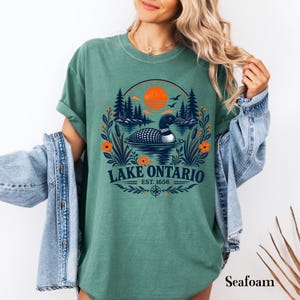May include: A seafoam green t-shirt featuring a graphic of a loon swimming on Lake Ontario. The design includes a sunset, pine trees, cattails, and orange flowers. The text reads Lake Ontario Est. 1656, framed by a circular border and compass.