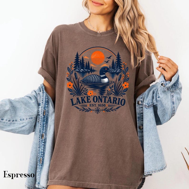 May include: An espresso-colored t-shirt featuring a graphic of a loon swimming on a lake, surrounded by pine trees, cattails, and orange flowers under an orange sun. The text reads LAKE ONTARIO EST. 1656 in dark blue lettering.