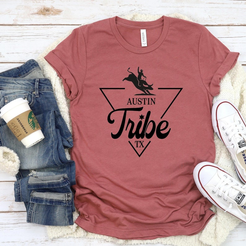 May include: A maroon t-shirt with a black graphic design of a cowboy riding a bull inside a triangle. The text "Austin Tribe TX" is below the graphic.