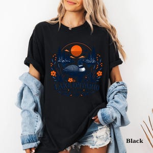 May include: A black t-shirt featuring a graphic print of a loon swimming on a lake, surrounded by pine trees and orange flowers under a setting sun. The text "LAKE ONTARIO" is printed in blue below the loon, with "EST. 1784" underneath.