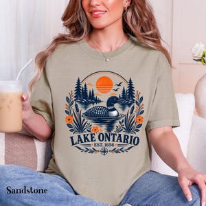 May include: A sandstone-colored t-shirt featuring a graphic of a loon swimming on a lake with pine trees, an orange sun, and floral accents. The text reads LAKE ONTARIO EST. 1656, accompanied by a compass rose icon at the bottom of the design.