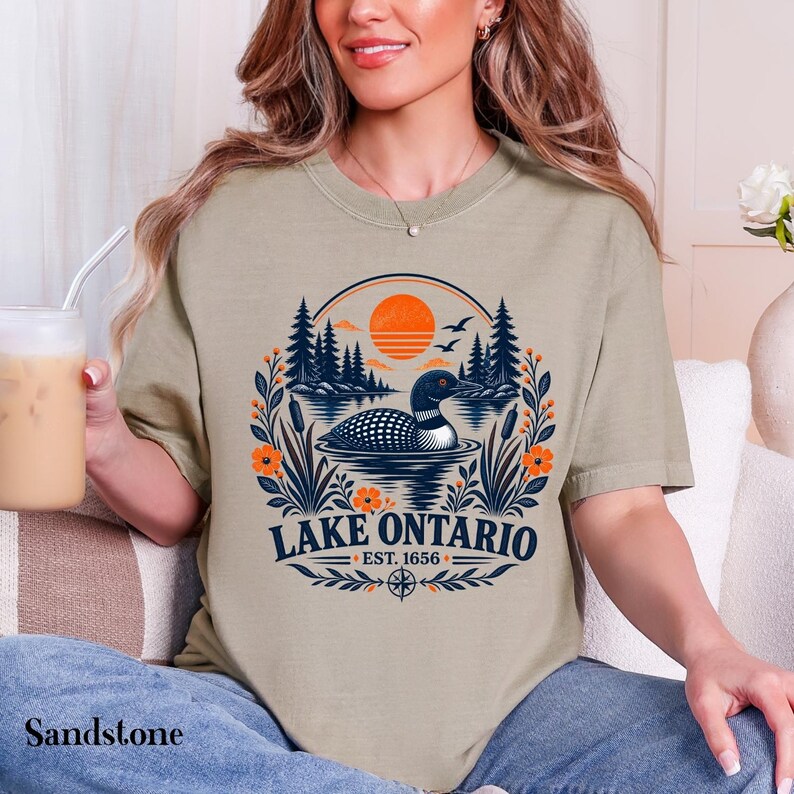 May include: A sandstone-colored t-shirt featuring a graphic of a loon swimming on a lake with pine trees, an orange sun, and floral accents. The text reads LAKE ONTARIO EST. 1656, accompanied by a compass rose icon at the bottom of the design.