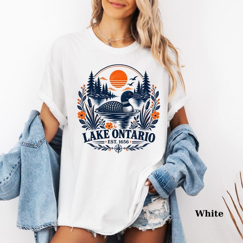May include: A white t-shirt featuring a graphic of a loon swimming on a lake with pine trees, orange sun, and floral accents. The text reads LAKE ONTARIO EST. 1656. The shirt is layered under a light blue denim jacket, creating a casual look.