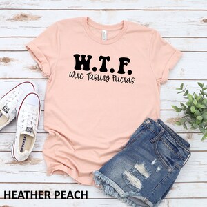 K&ouml;nnte beinhalten: Ein pfirsichfarbenes T-Shirt mit dem Text "W.T.F. Wine Tasting Friends" in Schwarz. Das Shirt liegt auf einer Holzoberfl&auml;che mit einem Paar wei&szlig;en Turnschuhen und einer Jeans-Shorts.