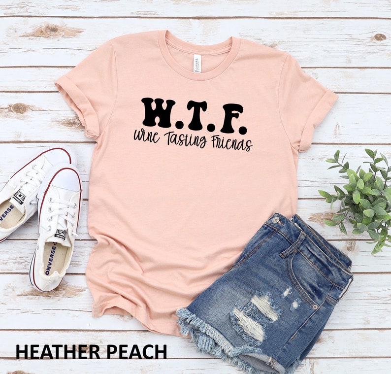 K&ouml;nnte beinhalten: Ein pfirsichfarbenes T-Shirt mit dem Text "W.T.F. Wine Tasting Friends" in Schwarz. Das Shirt liegt auf einer Holzoberfl&auml;che mit einem Paar wei&szlig;en Turnschuhen und einer Jeans-Shorts.