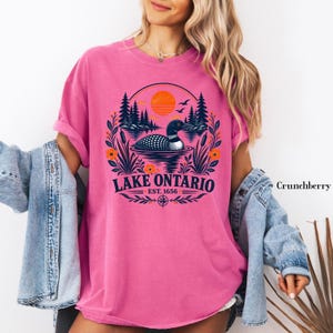 May include: A bright pink short-sleeve t-shirt featuring a graphic of a loon swimming on a lake with pine trees, cattails, and an orange sun. The text reads Lake Ontario Est. 1656. A light blue denim jacket is layered over the pink t-shirt.