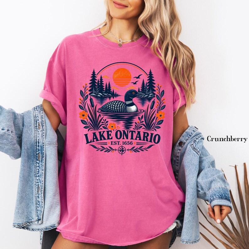May include: A bright pink short-sleeve t-shirt featuring a graphic of a loon swimming on a lake with pine trees, cattails, and an orange sun. The text reads Lake Ontario Est. 1656. A light blue denim jacket is layered over the pink t-shirt.
