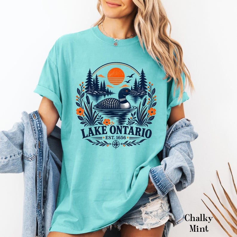 May include: A mint green short-sleeve t-shirt featuring a graphic of a loon swimming on a lake with pine trees, cattails, and an orange sun. The text reads LAKE ONTARIO EST. 1656, framed by floral elements and a compass rose at the bottom.