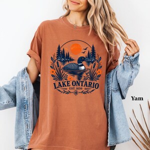 May include: A rust-colored t-shirt featuring a graphic of a loon swimming on Lake Ontario. The design includes a sunset, pine trees, cattails, and the text "LAKE ONTARIO EST. 1656" in dark blue. The shirt is worn with a light blue denim jacket.