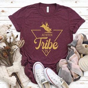 May include: A maroon t-shirt with a gold graphic of a bull rider in a triangle with the text "Austin Tribe TX".