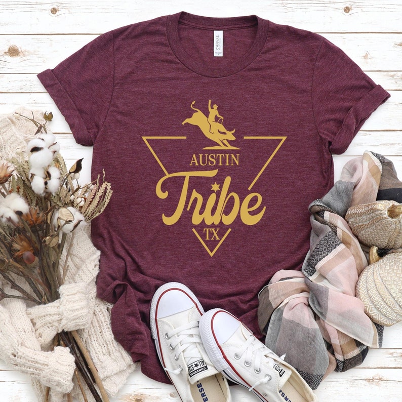 May include: A maroon t-shirt with a gold graphic of a bull rider in a triangle with the text "Austin Tribe TX".
