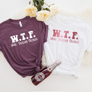 K&ouml;nnte beinhalten: Zwei T-Shirts mit dem Text "W.T.F. Wine Tasting Friends" in einer Retro-Schriftart. Ein Shirt ist wei&szlig; mit rosa Schrift und das andere ist lila mit wei&szlig;er Schrift. Eine Flasche Sekt befindet sich in der Mitte des Bildes.