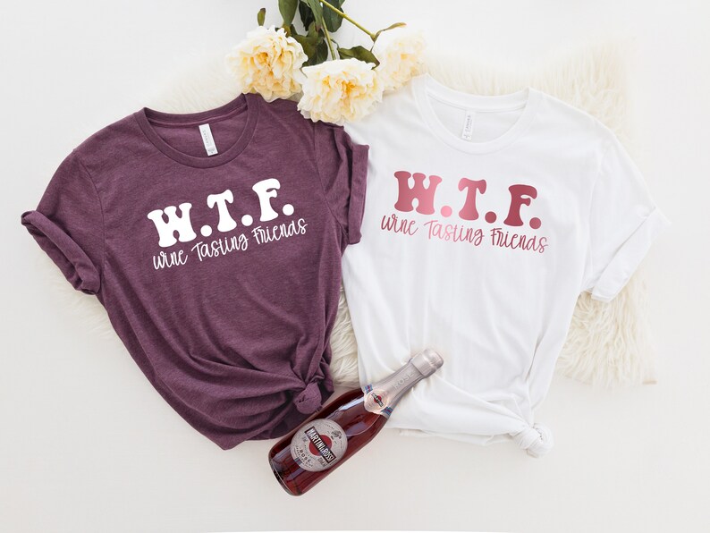 K&ouml;nnte beinhalten: Zwei T-Shirts mit dem Text "W.T.F. Wine Tasting Friends" in einer Retro-Schriftart. Ein Shirt ist wei&szlig; mit rosa Schrift und das andere ist lila mit wei&szlig;er Schrift. Eine Flasche Sekt befindet sich in der Mitte des Bildes.