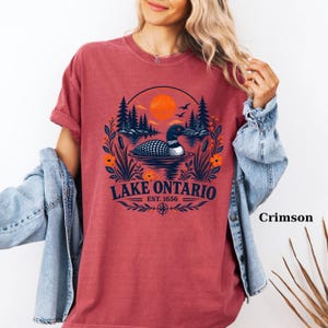 May include: A crimson short-sleeve t-shirt featuring a graphic of a loon swimming on a lake with pine trees, cattails, and an orange sun. The text reads LAKE ONTARIO EST. 1656. The shirt is worn with a light blue denim jacket over the shoulders.