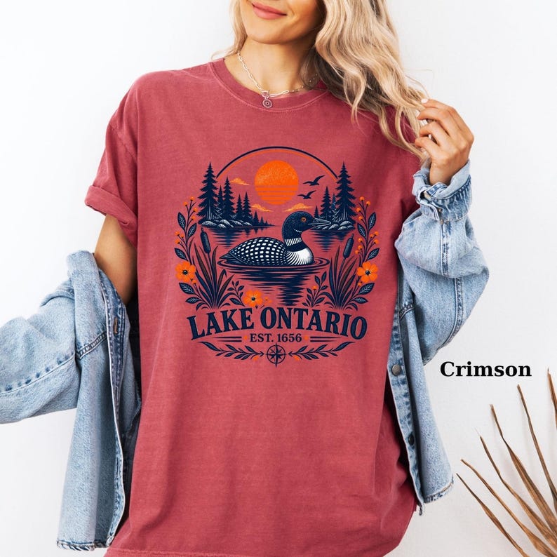 May include: A crimson short-sleeve t-shirt featuring a graphic of a loon swimming on a lake with pine trees, cattails, and an orange sun. The text reads LAKE ONTARIO EST. 1656. The shirt is worn with a light blue denim jacket over the shoulders.