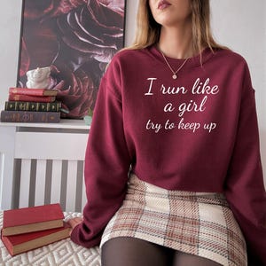 May include: Burgundy sweatshirt with the text "I run like a girl try to keep up" in white. The sweatshirt is paired with a plaid skirt. A gold necklace is visible. The image is of a person.