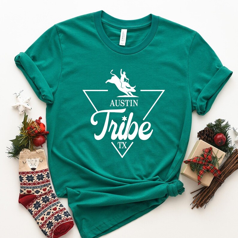 May include: A teal green t-shirt with a white graphic design. The design features a silhouette of a cowboy riding a bull, a triangle, and the text "Austin Tribe TX".