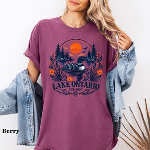 May include: A berry-colored short-sleeve t-shirt featuring a graphic of a loon swimming on a lake with pine trees, an orange sun, and flowers. The text reads Lake Ontario Est. 1656. The design includes a compass rose and foliage elements.
