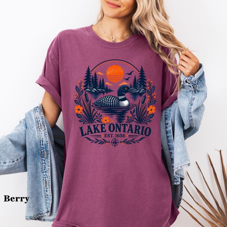 May include: A berry-colored short-sleeve t-shirt featuring a graphic of a loon swimming on a lake with pine trees, an orange sun, and flowers. The text reads Lake Ontario Est. 1656. The design includes a compass rose and foliage elements.