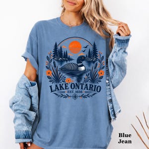 May include: A blue t-shirt featuring a graphic of a loon swimming on a lake with pine trees, cattails, and an orange sun. The text reads LAKE ONTARIO EST. 1656. The shirt is worn with a blue denim jacket, creating a layered look with a casual style.