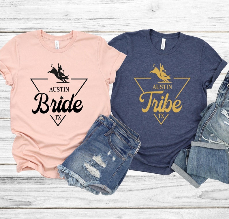 May include: Two t-shirts with a graphic design of a cowboy riding a bull. The pink shirt says "Austin Bride TX" and the blue shirt says "Austin Tribe TX".