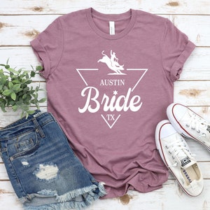 May include: A mauve t-shirt with a white graphic of a bull rider in a triangle with the text "Austin Bride TX".