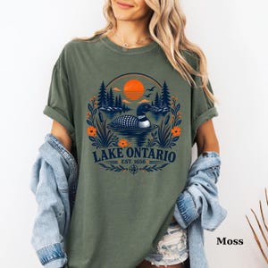 May include: A moss green t-shirt featuring a graphic of a loon swimming on a lake, surrounded by pine trees, orange flowers, and a sunset. The text reads LAKE ONTARIO EST. 1656 in dark blue lettering, with a compass rose icon below the text.