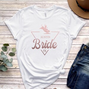 May include: White t-shirt with a rose gold graphic of a bull rider in a triangle with the text "Austin Bride TX"