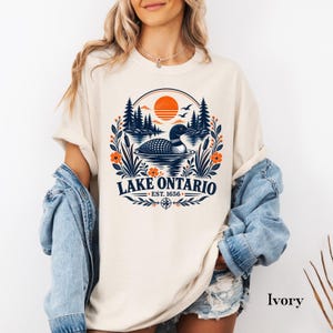 May include: An ivory t-shirt featuring a graphic print of a loon swimming on Lake Ontario. The design includes a sunset, pine trees, cattails, and orange flowers, with the text LAKE ONTARIO EST. 1656 centered below the circular nature scene.