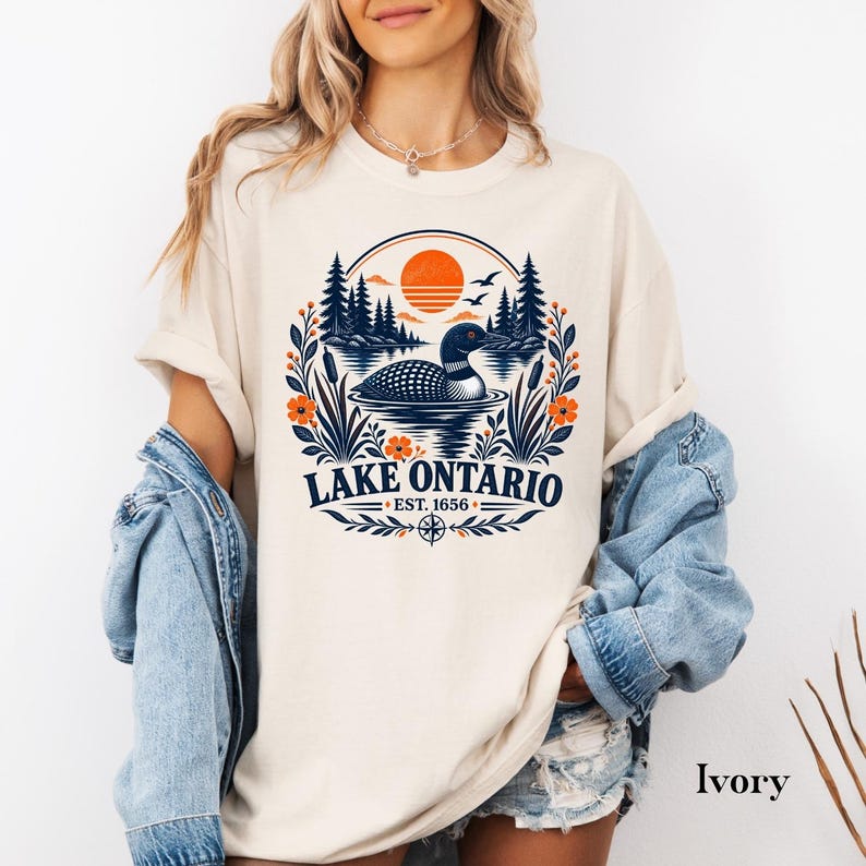 May include: An ivory t-shirt featuring a graphic print of a loon swimming on Lake Ontario. The design includes a sunset, pine trees, cattails, and orange flowers, with the text LAKE ONTARIO EST. 1656 centered below the circular nature scene.