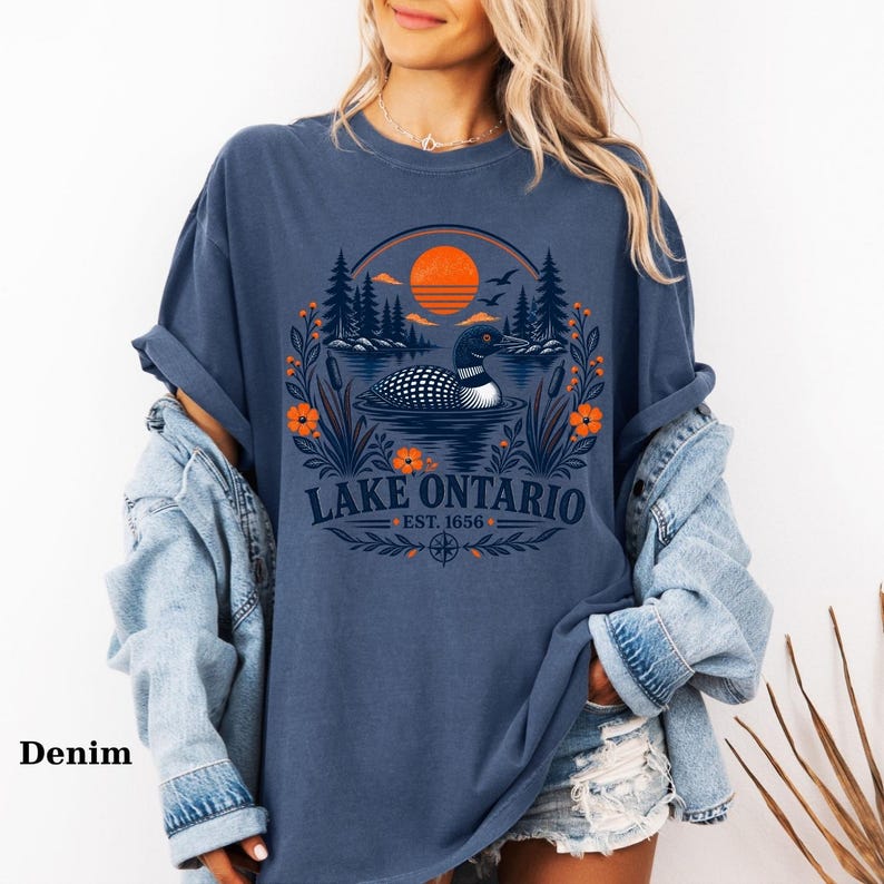 May include: A denim blue t-shirt featuring a graphic print of a loon swimming on a lake with pine trees, orange flowers, and a sunset. The text reads Lake Ontario Est. 1656. The shirt is worn with a light blue denim jacket and distressed denim shorts.