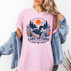 May include: A light pink short-sleeve t-shirt featuring a graphic of a loon swimming on a lake with pine trees and an orange sun. The text reads Lake Ontario Est. 1656, surrounded by a circular floral border with cattails and orange flowers.