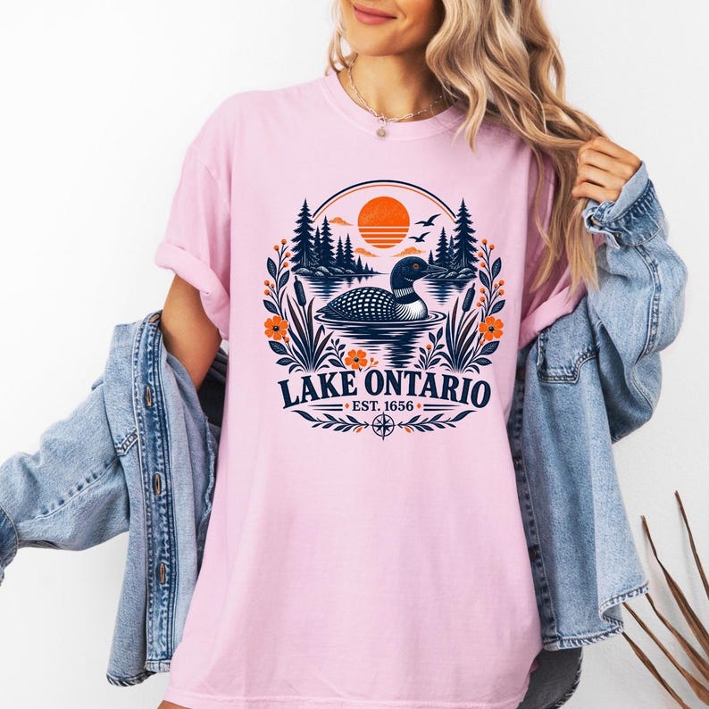May include: A light pink short-sleeve t-shirt featuring a graphic of a loon swimming on a lake with pine trees and an orange sun. The text reads Lake Ontario Est. 1656, surrounded by a circular floral border with cattails and orange flowers.