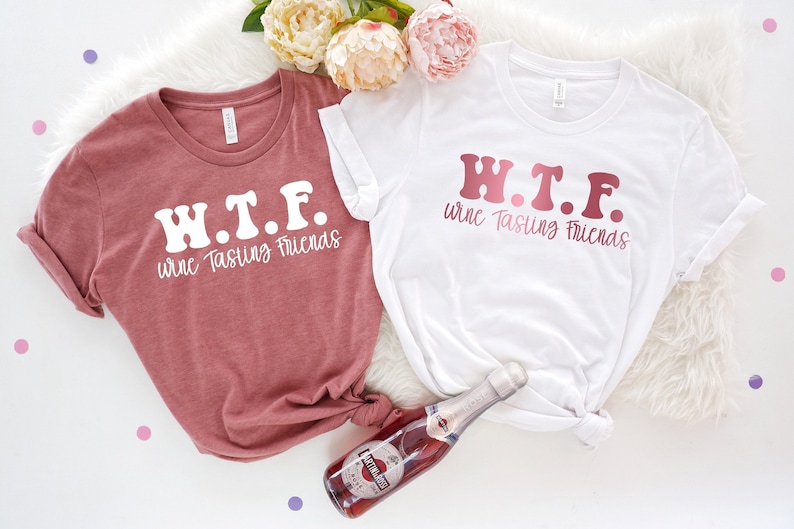 K&ouml;nnte beinhalten: Zwei wei&szlig;e T-Shirts mit dem Text "W.T.F. wine Tasting Friends" in rosa und wei&szlig;er Schrift. Die Shirts liegen auf einer wei&szlig;en, flauschigen Oberfl&auml;che mit einer Flasche rosafarbenem Sekt.