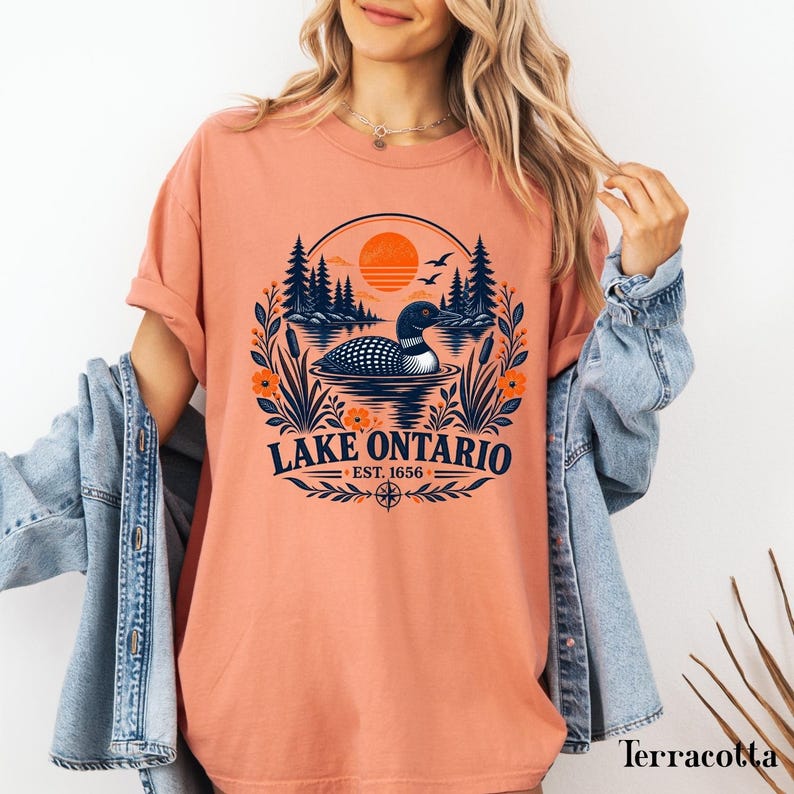 May include: A terracotta-colored t-shirt featuring a graphic print of a loon swimming on a lake, surrounded by pine trees, cattails, and flowers. The text reads Lake Ontario Est. 1656, accompanied by a sun, birds, and a compass rose design.