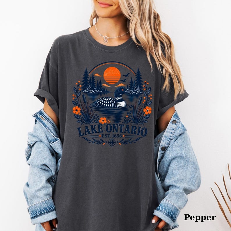 May include: A charcoal gray t-shirt featuring a graphic of a loon swimming on a lake with pine trees and an orange sun. The text reads Lake Ontario Est. 1656, surrounded by floral elements and a compass rose design at the bottom of the print.