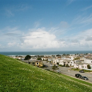May include: A scenic view of a coastal town under a bright blue sky with scattered clouds. A grassy hill slopes down towards a road lined with buildings and cars, with the ocean visible in the distance.