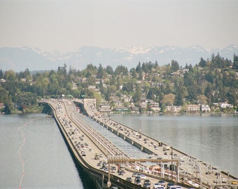 May include: An aerial view of a bridge over a body of water, with cars driving in both directions. The bridge is surrounded by lush green trees and houses on a hillside. Mountains are visible in the background.