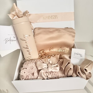 May include: A white gift box filled with a variety of items for a bridesmaid. The items include a personalised tumbler with a straw, a satin eye mask, a hair clip, a scrunchie, and a lace headband. The box is decorated with a ribbon and the name "Kimberly" is printed on the ribbon.