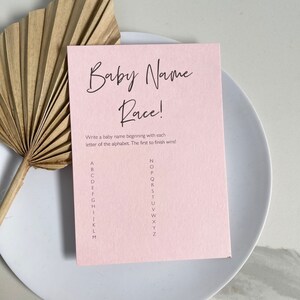 May include: A pink card with the text "Baby Name Race!" and instructions to write a baby name beginning with each letter of the alphabet. The first to finish wins! The alphabet is listed in two columns.