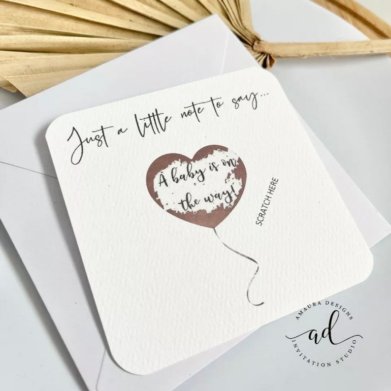 Puede incluir: Una tarjeta blanca con el texto "Just a little note to say... A baby is on the way!" El texto est&aacute; escrito en letra cursiva negra. Un globo en forma de coraz&oacute;n marr&oacute;n con una cuerda blanca est&aacute; en el centro de la tarjeta. El texto "Scratch Here" est&aacute; escrito en negro en el lado derecho de la tarjeta. La parte inferior de la tarjeta tiene el texto "Amaura Designs" y "Invitation Studio" en letra cursiva negra.
