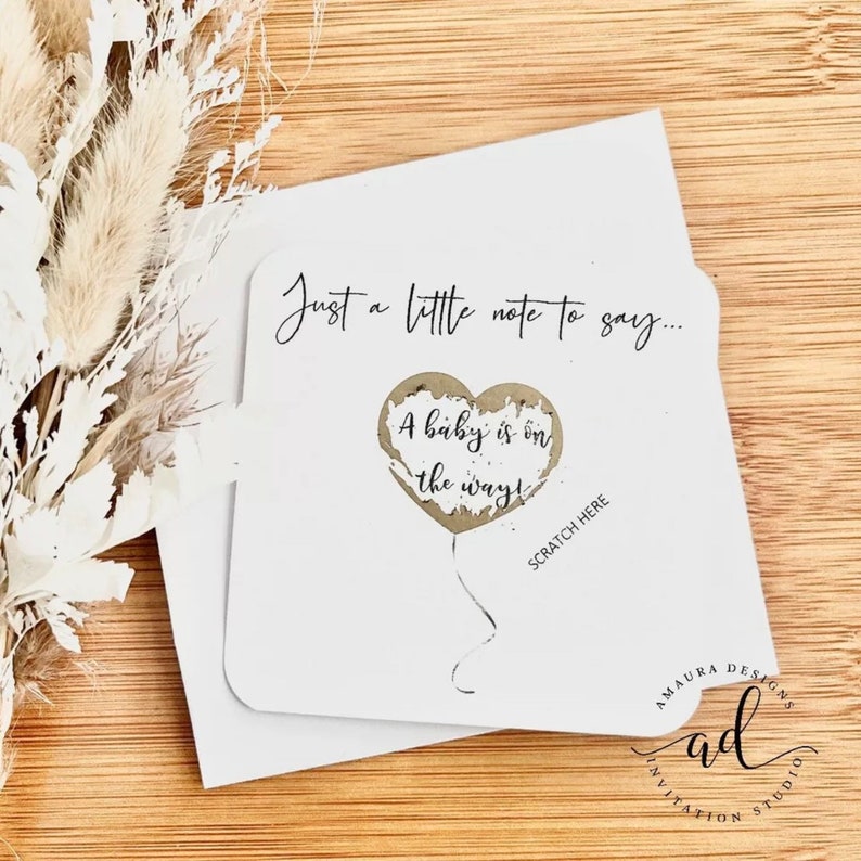 Puede incluir: Una tarjeta blanca con el texto "Just a little note to say... A baby is on the way!" con un gr&aacute;fico de un globo en forma de coraz&oacute;n dorado. El texto "Scratch Here" est&aacute; debajo del globo. La tarjeta est&aacute; sobre una superficie de madera.