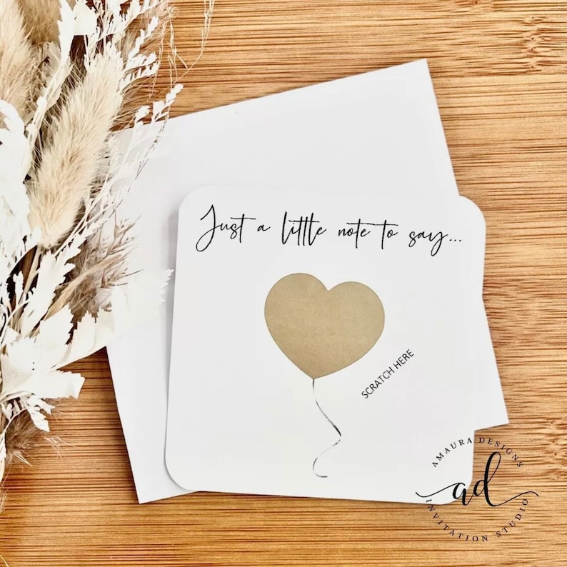 Puede incluir: Una tarjeta blanca con el texto "Just a little note to say..." y un globo en forma de coraz&oacute;n con un &aacute;rea para rascar que dice "Scratch Here". La tarjeta es de Amaura Designs.