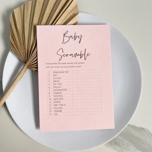 May include: A pink printable baby shower game card with the title "Baby Scramble". The game instructions are to unscramble the baby words and the person with the most correct answers wins. The game includes 15 words to unscramble.