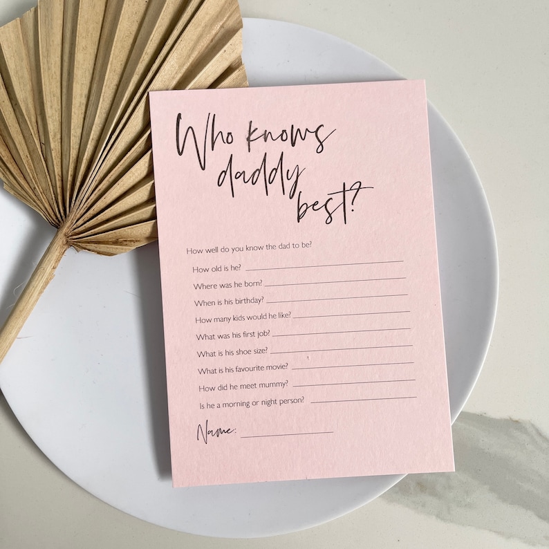 May include: A pink printable game card with the text "Who knows daddy best?" and a list of questions about a father. The card is on a white plate with a brown fan behind it.